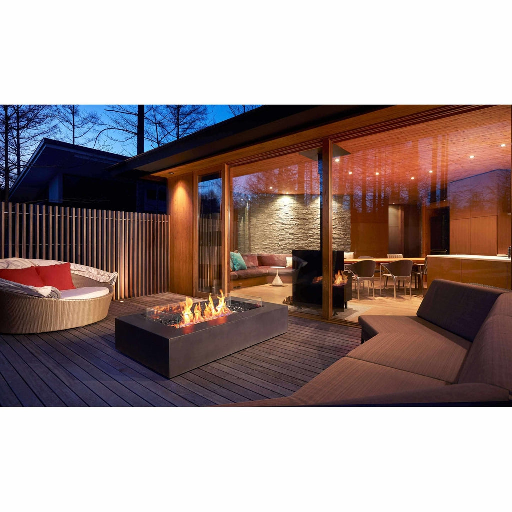 EcoSmart Fire Wharf 65 ethanol fire pit table with glass screen on a dark wooden deck at twilight.
