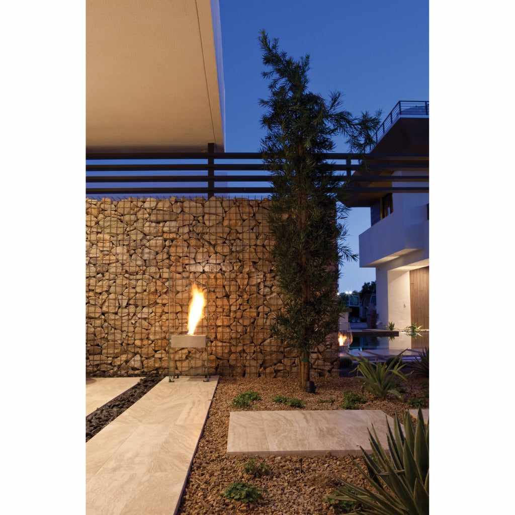 EcoSmart Fire Ghost bioethanol fireplace with a clear glass frame burning on a patio against a stone gabion wall.