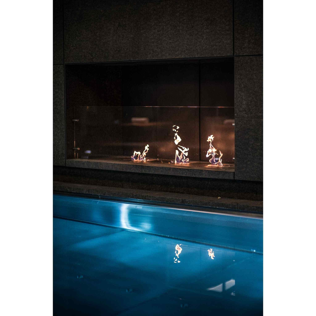 EcoSmart Fire BK5 ethanol burner with flames behind a glass screen, set into a dark stone wall above a blue pool.