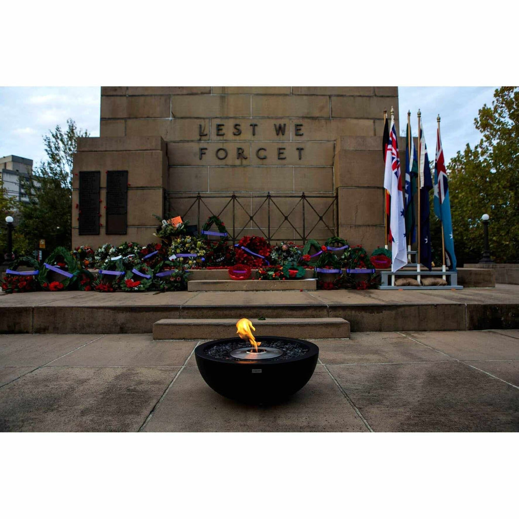 EcoSmart Fire Mix 600 bioethanol fire pit bowl with a lit flame, placed on a stone plaza before a war memorial.