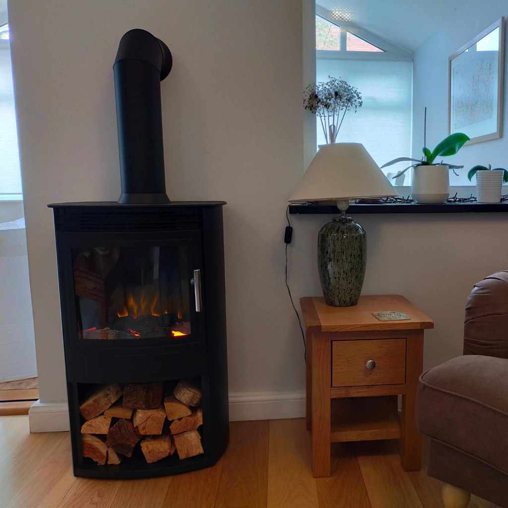 Flametek Alto T electric stove in black with a log storage compartment, placed on a wooden floor next to a side table.