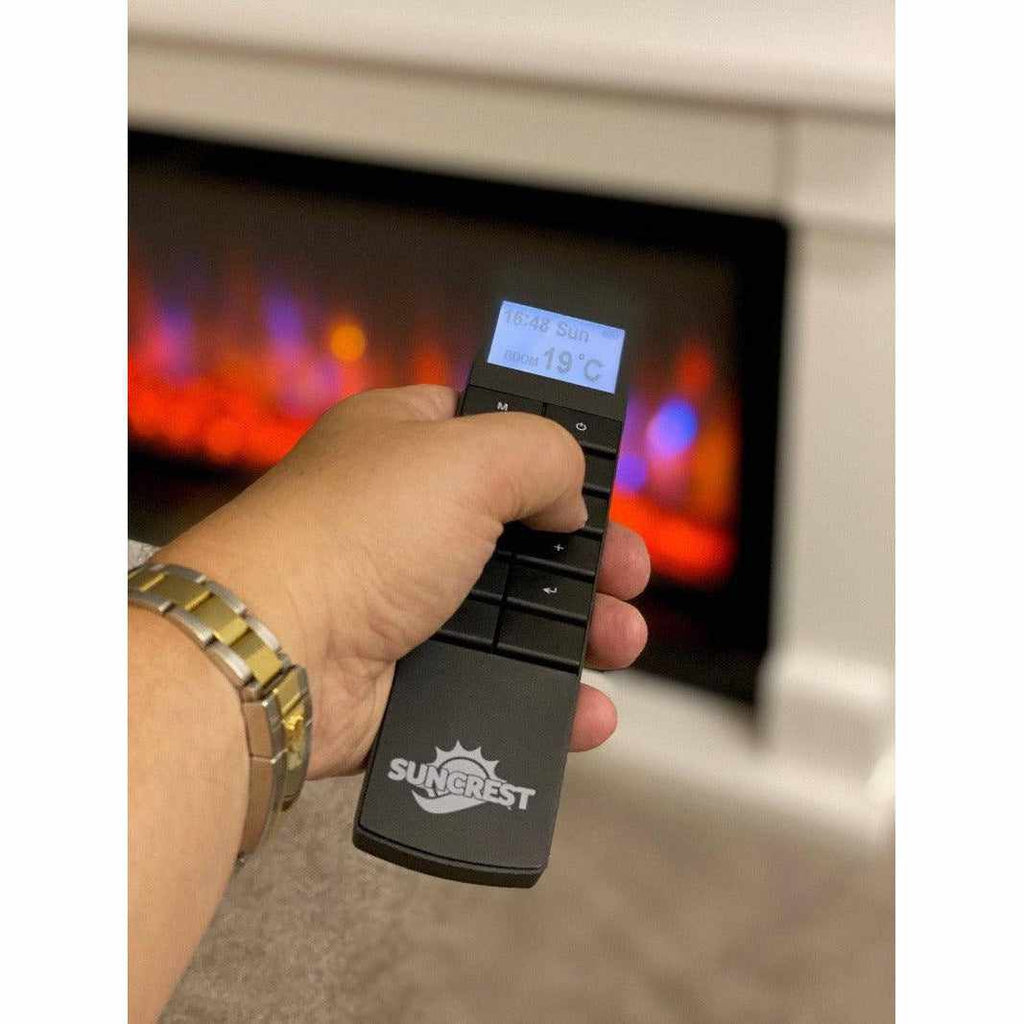 Hand holding a black Suncrest Bradbury electric fireplace remote with an illuminated digital display.