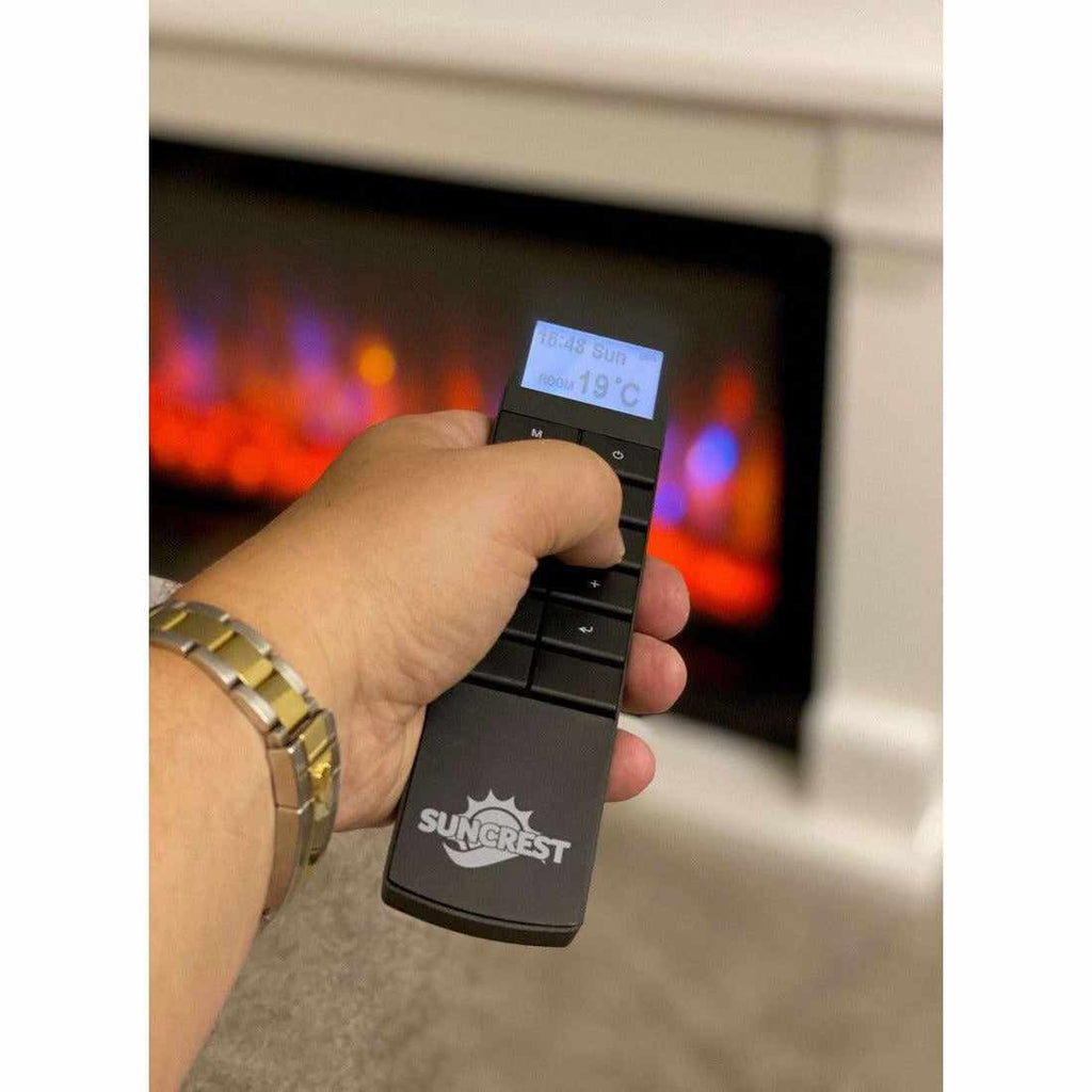 Hand holding a black Suncrest Lumley electric fireplace remote with an illuminated digital display.